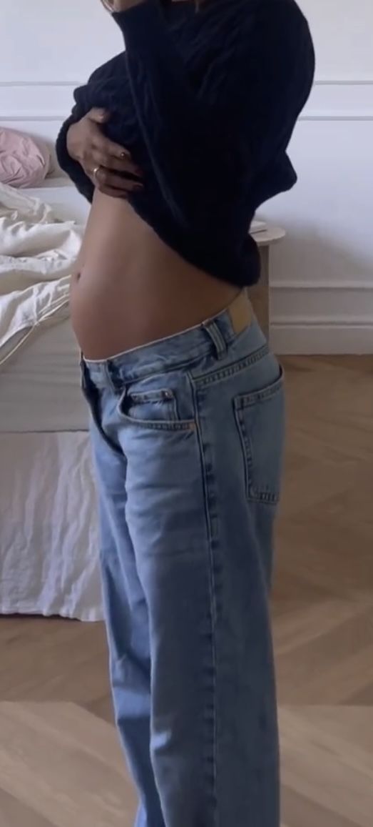 a pregnant woman is standing in her jeans and looking down at her stomach, with one hand on her hip