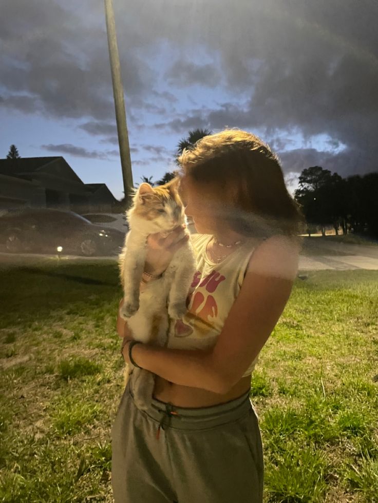 a woman holding a cat while standing on top of a lush green field under a cloudy sky