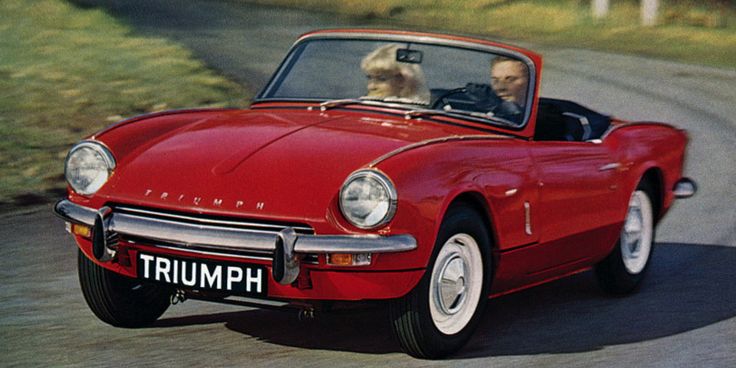 an old red triumph sports car driving down a road with two people in the back seat