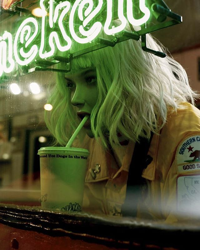 a woman with green hair is drinking from a cup in front of a neon sign