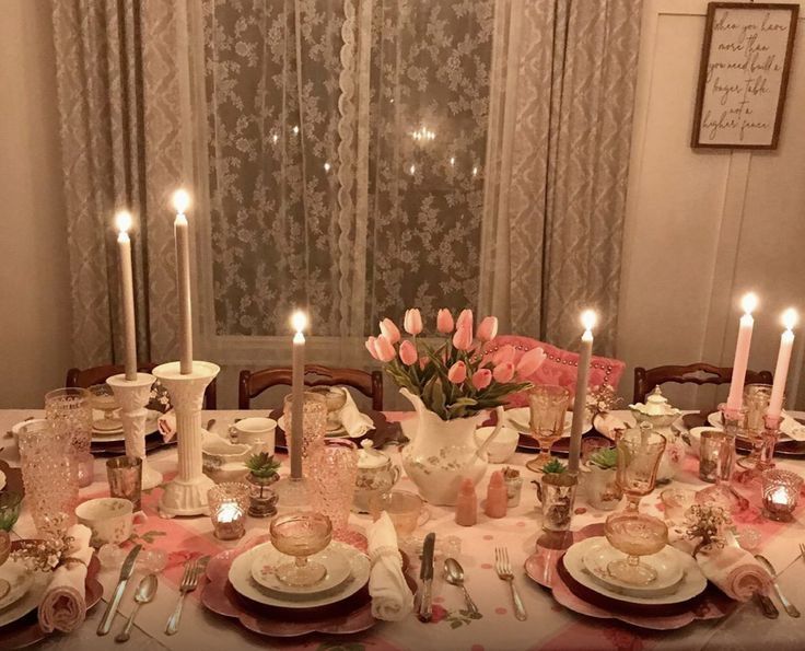 a dining room table is set with pink flowers and white china plates, candles, and napkins