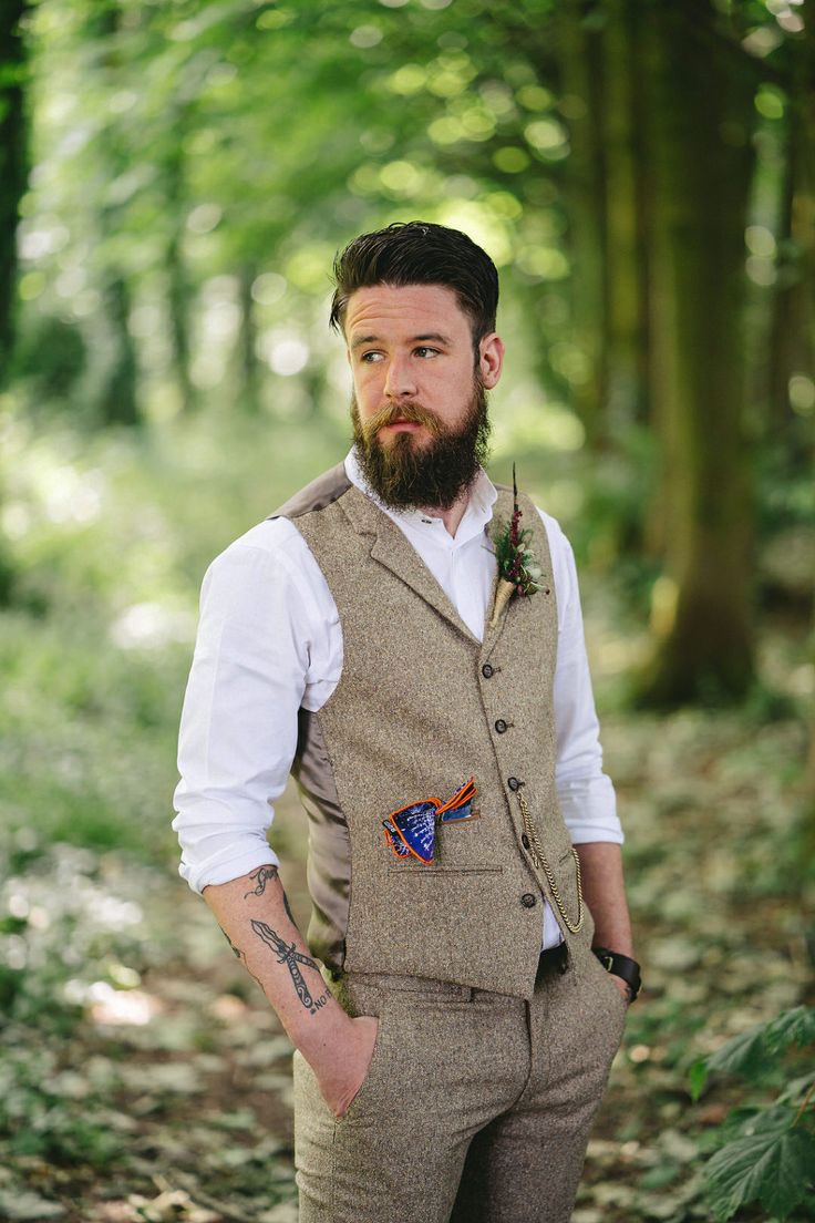 a man with a beard wearing a vest and bow tie standing in the middle of a forest