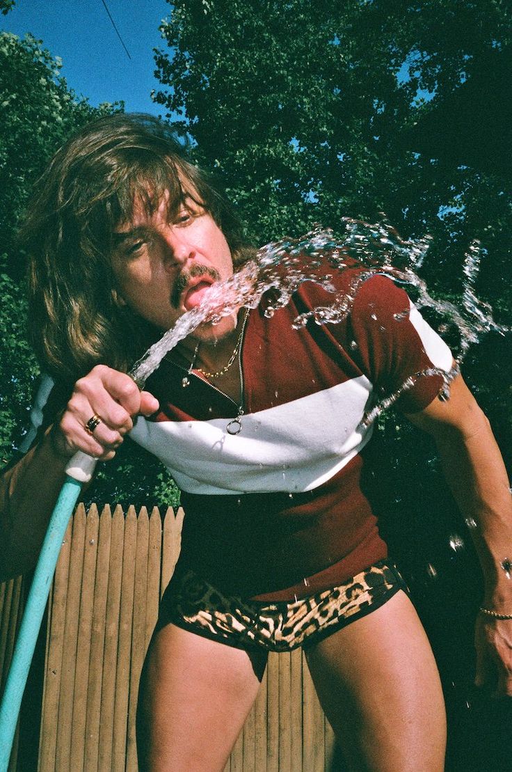 a man with long hair is spraying water on his face and holding a blue hose