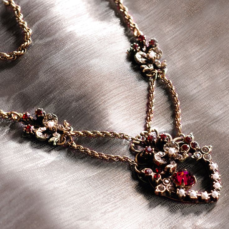 "Necklace: The festoon style was introduced in the early 1900s and this particular theme was given and received by both friends and those with romantic interests. A floral and bow motif, this design is set with garnet and smoke crystal and seed pearls An oval faceted Czech garnet stone dangles inside the heart. An oval prong set stone dangles separately inside the heart. Vintage double twist rope chain connects the pendant and side ornaments. Metal finish is burnished bronze. Necklace Measuremen Heirloom Necklace With Vintage Charm For Valentine's Day, Elegant Vintage Heart Pendant Jewelry, Vintage Jewelry For Celebration With Jewels, Victorian Necklace With Historical Design As Gift, Vintage Jewelry With Historical Design, Red Victorian Necklace For Anniversary, Vintage Jewelry With Intricate Design For Celebrations, Vintage Jewelry For Celebration With Intricate Design, Elegant Necklaces With Intricate Design For Valentine's Day