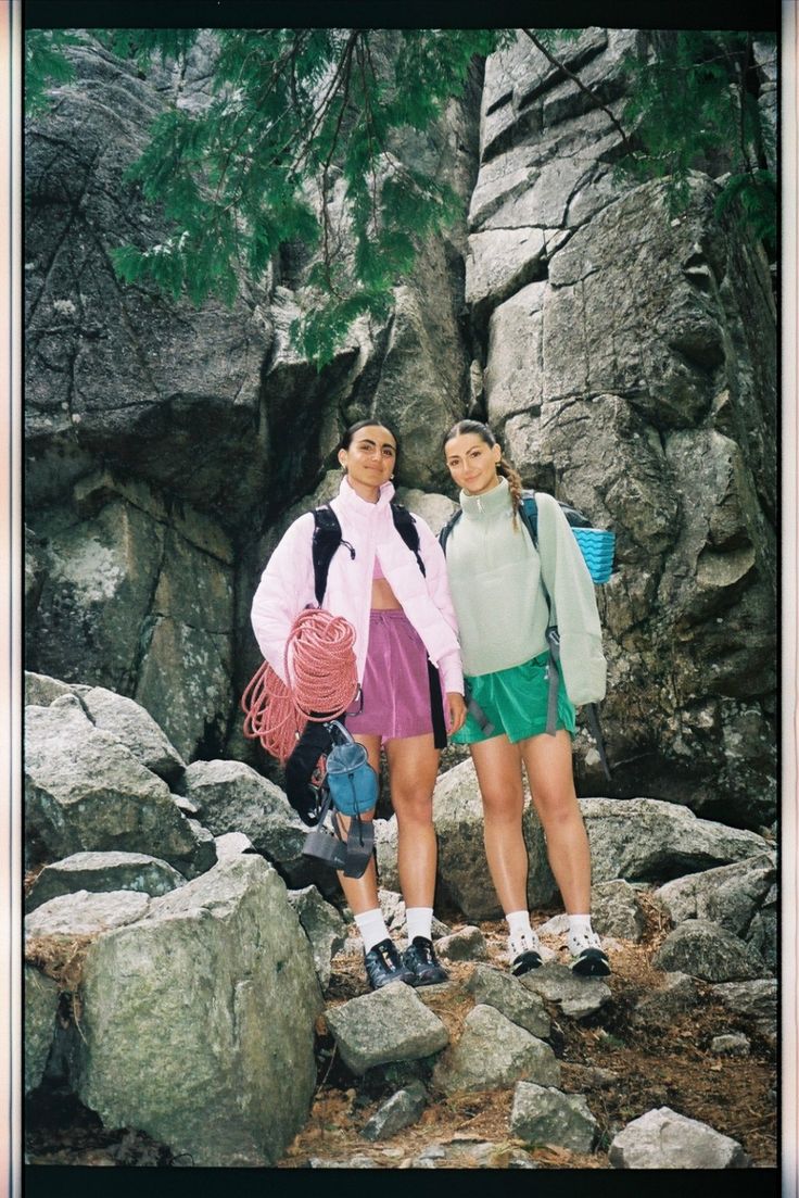 two women standing next to each other in front of some rocks and trees with their backpacks