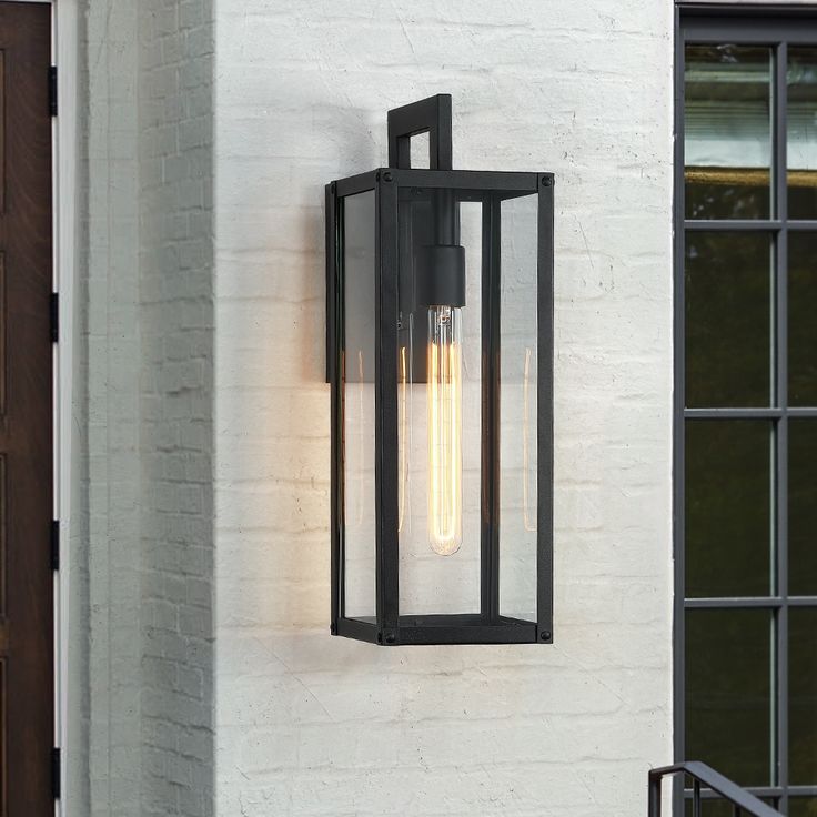 Illuminate your front door or garage with this contemporary exterior wall mounted lantern light. It's rated for wet locations, and its sleek frames are made from metal in a powder-coated black finish that complements your decor from classic to rustic. It features 4 clear glass panels with a elongated shape enclosed in a black and sleek clean-lined framework. E26 long tubular bulb is recommended to fully illuminate the glass and to complement the overall aesthetic, or use a Edison bulb to enhance a vintage look. It's Ideal for Front Porch, Patio, Garage, Deck etc. Plus, it's compatible with a dimmer switch, so you can make the most out of your lighting. True Fine Spun 1-Light 18.5-in H Matte Black LED Outdoor Wall Light | TD130013OT Black Outdoor Lights, House Lighting Outdoor, Outdoor Wall Light Fixtures, Wall Mount Lantern, Exterior Light Fixtures, Black Outdoor Wall Lights, Contemporary Exterior, Led Outdoor Wall Lights, Garage Lighting