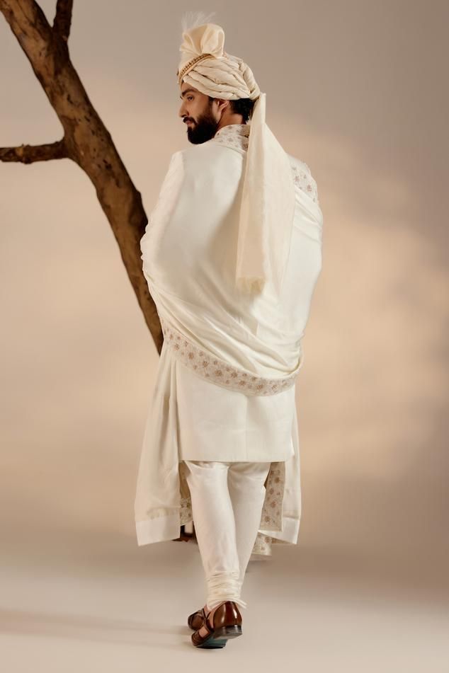 White full sleeves sherwani in linen silk base with all over Kashmiri Jamawar embroidery highlighted wih dabka and zari. Paired with full sleeves plain kurta, churidar, safa and embroidered border stole.
Components: 5
Pattern: Embroidery
Type Of Work: Zari, Dabka
Neckline: Band
Sleeve Type: Long
Fabric: Linen Silk
Color: White
Other Details: 
Closure: 
Sherwani: Front hooks
Kurta: Front placket buttons
Note: The jewellery and kalangi worn by the model is not for sale
Occasion: Groom - Aza Fashio Cotton Silk Sherwani With Chikankari Embroidery, White Cotton Silk Sherwani With Long Sleeves, Ceremonial Cotton Silk Sherwani With Resham Embroidery, White Cotton Silk Long Sleeve Sherwani, Traditional Wear With Naqshi On Cotton Silk, Traditional Wear With Naqshi In Cotton Silk, Naqshi Cotton Silk Traditional Wear, White Cotton Silk Sherwani With Dabka, Cotton Silk Sherwani With Cutdana In Traditional Drape