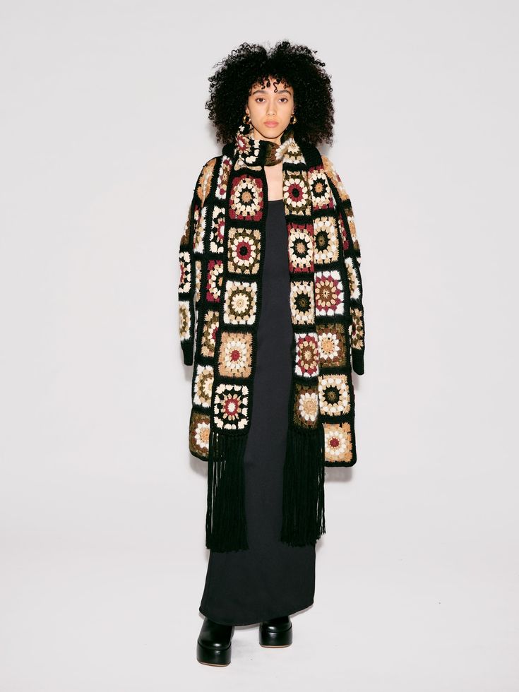 a woman standing in front of a white wall wearing a black dress and a crocheted coat