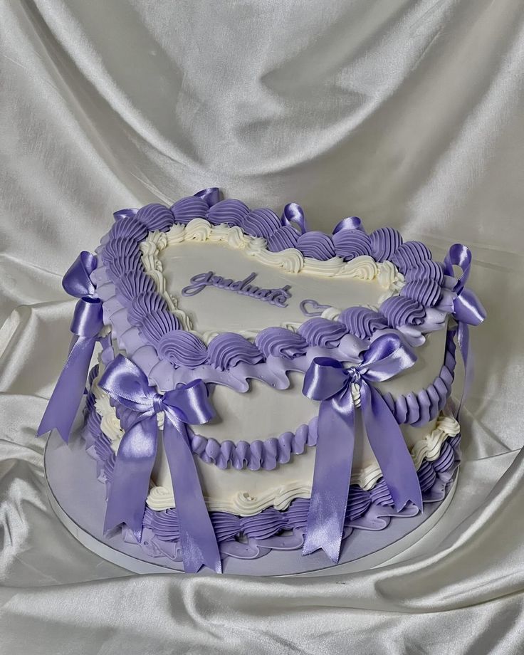 a heart shaped cake with purple ribbons and bows on the top is sitting on a silver cloth