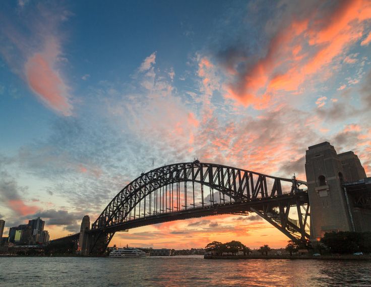 the sydney harbour bridge at sunset as seen from across the water with clouds in the sky