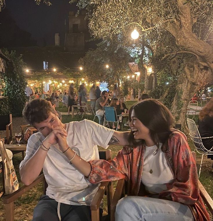 two people sitting on wooden chairs in an outdoor area at night, one holding his face to the other's ear