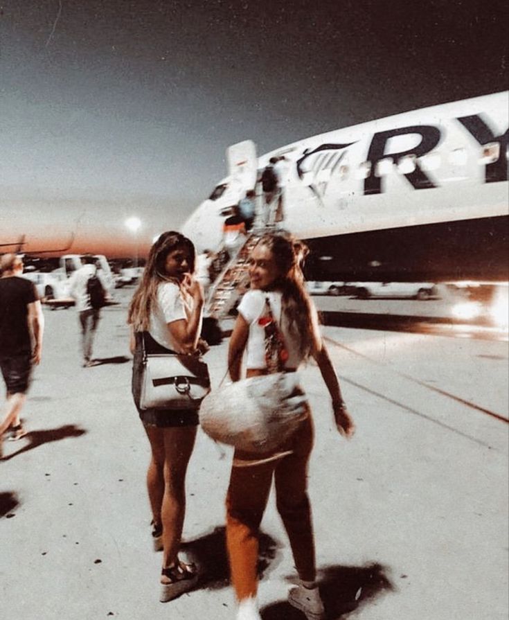 two women are walking towards an airplane on the tarmac at night, with other people in the background