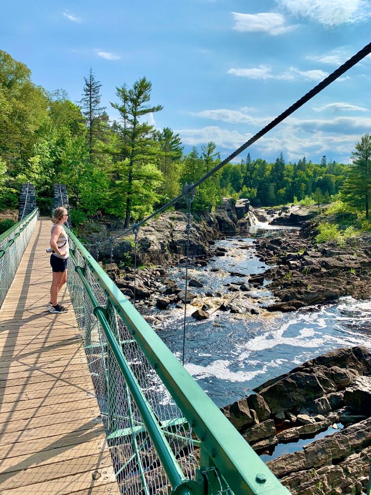 a woman walking across a bridge over a river next to a lush green forest filled with trees