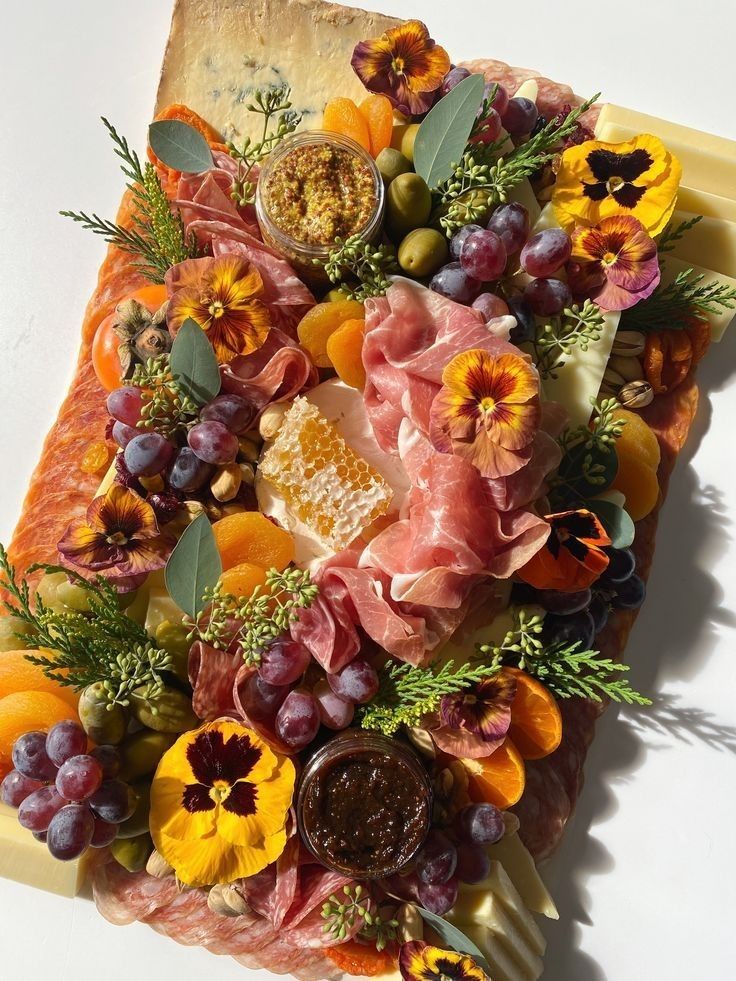 an arrangement of fruits, meats and flowers on top of a piece of cheese