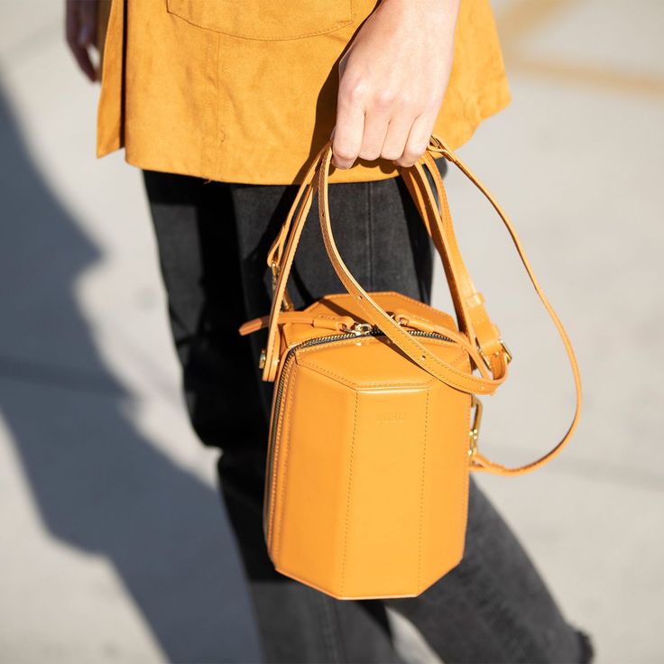 The cute Harper unexpected geometric shape gives this orange leather crossbody bag a playful but sophisticated runway savvy look. With its gold hardware, its small handle allows this orange leather bucket bag to swivel and be held in hand as a bucket. Its top zipper will keep your contents secure inside, while the cotton lined interior with 3 compartments keeps everything organized. This trendy designer crossover barrel bucket purse comes with a long and comfortable leather strap that can be flu Shopping Bucket Bag With Zipper Closure, Modern Bucket Bag With Zipper Closure, Orange Crossbody Bucket Bag, Modern Handheld Bucket Bag With Gold-tone Hardware, Orange Bucket Bag For Everyday Use, Orange Bucket Bag With Adjustable Strap, Orange Bucket Bag For Daily Use, Chic Orange Bucket Bag Tote, Modern Bucket Style Box Bag For Travel