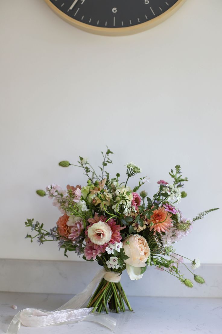 Natural style bridal bouquet in peach and pink with fresh green foliage and organza ribbons sits on a marble side. Simple Wedding Bouquet Colorful, Bride Bouquet Flower Types, March Wedding Flowers Uk, Garden Flowers Wedding Bouquet, Wedding Summer Florals, English Wedding Bouquet, Summer Bridal Bouquet Romantic, English Garden Bridal Bouquet, Wedding Flowers Romantic