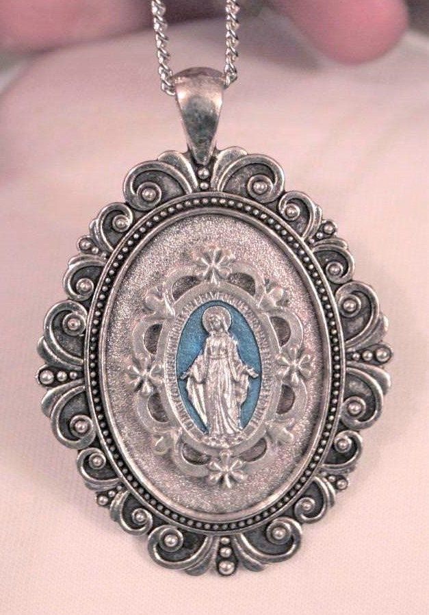 This silvertone necklace features a lovely small-to-mid-sized pendant with a swoop scalloped outer rim that gives it charm and character.  The center is accented with a blue finished floral medal inset with the image of The Immaculate Conception, Our Lady of Grace -  The Miraculous Mary.  It is inscribed with the Miraculous Prayer and the pendant has a shiny finish on the reverse.  It measures 1 5/8 inches wide and 2 3/8 inches long.  The chain is 24 inches and closes with a safety catch. Silver Necklaces With Oval Pendant Vintage Charm, Silver Necklace With Oval Pendant And Vintage Charm, Antique Silver Medallion With Miraculous Medal, Vintage Silver Necklace With Miraculous Medal, Vintage Miraculous Medal Jewelry, Our Lady Of Grace, Cross Medal, Lady Of Lourdes, Our Lady Of Lourdes