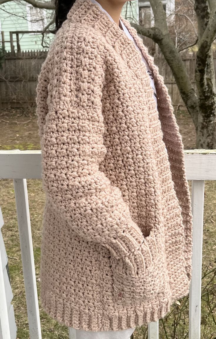 a woman standing on a porch wearing a pink crochet sweater and white pants