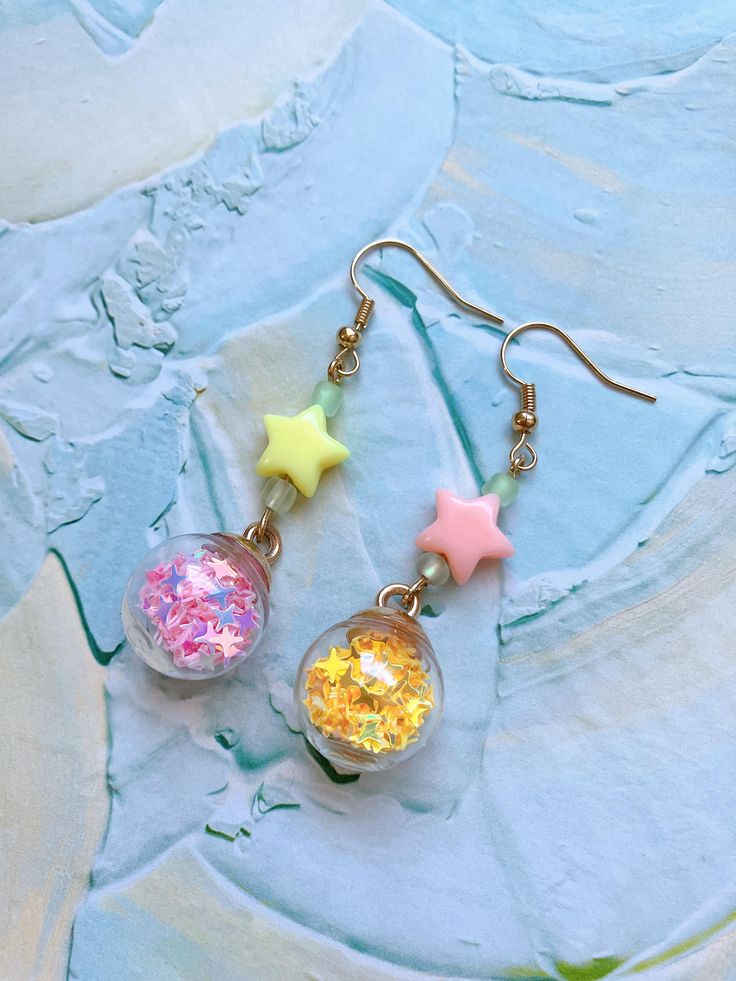 dd a touch of the night sky to your look with these enchanting star-themed dangle earrings. Each earring features a sparkling dome filled with tiny, twinkling stars, capturing the magic of a starlit night. Choose from two mesmerizing color pairs: ⭐︎Blue and Purple, reminiscent of a serene twilight sky, ⭐︎Yellow and Pink, evoking the glow of stars in a clear night sky. These lightweight plastic earrings are perfect for anyone who loves to carry a piece of the cosmos wherever they go. Whether you're dressing up for a special occasion or adding a bit of sparkle to your everyday outfit, these earrings are sure to catch the eye and brighten your day. Celestial Star-shaped Sparkling Earrings, Sparkling Star-shaped Celestial Earrings, Party Drop Earrings With Star Charm, Dangle Earrings With Star Charm For Party, Whimsical Star-shaped Jewelry With Star Charm, Whimsical Star-shaped Party Jewelry, Pink Star Charm Dangle Earrings, Pink Dangle Earrings With Star Charm, Star Charm Earrings For Party