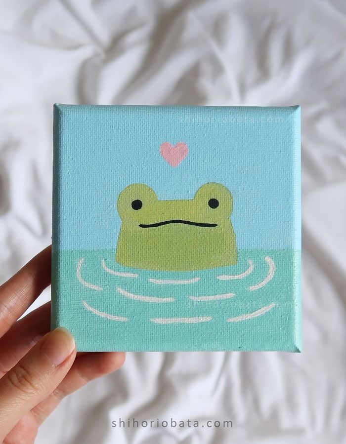 a hand holding up a small square painting with a frog in the water