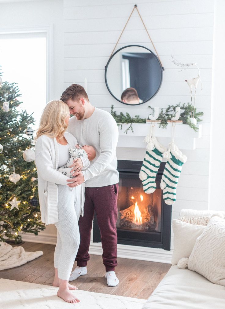 a man and woman standing in front of a christmas tree with stockings on the fireplace