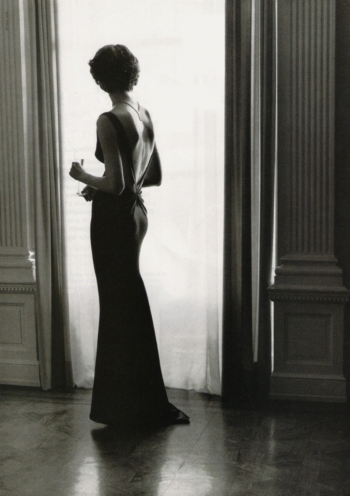 black and white photograph of woman in evening dress looking out the window with her back to the camera