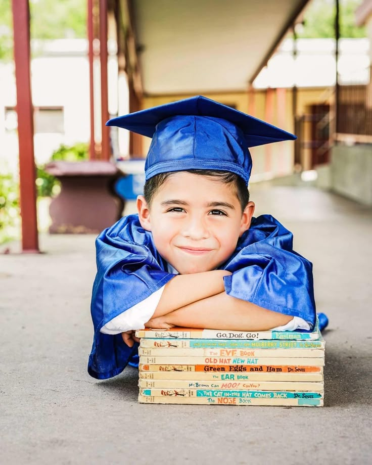 a young boy wearing a blue graduation cap and gown laying on top of stack of books