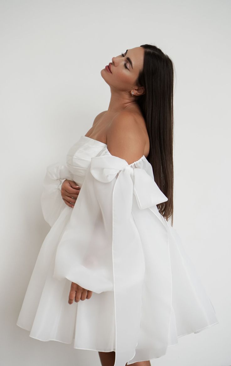 a woman with long hair wearing a white off the shoulder dress and holding her hands on her chest