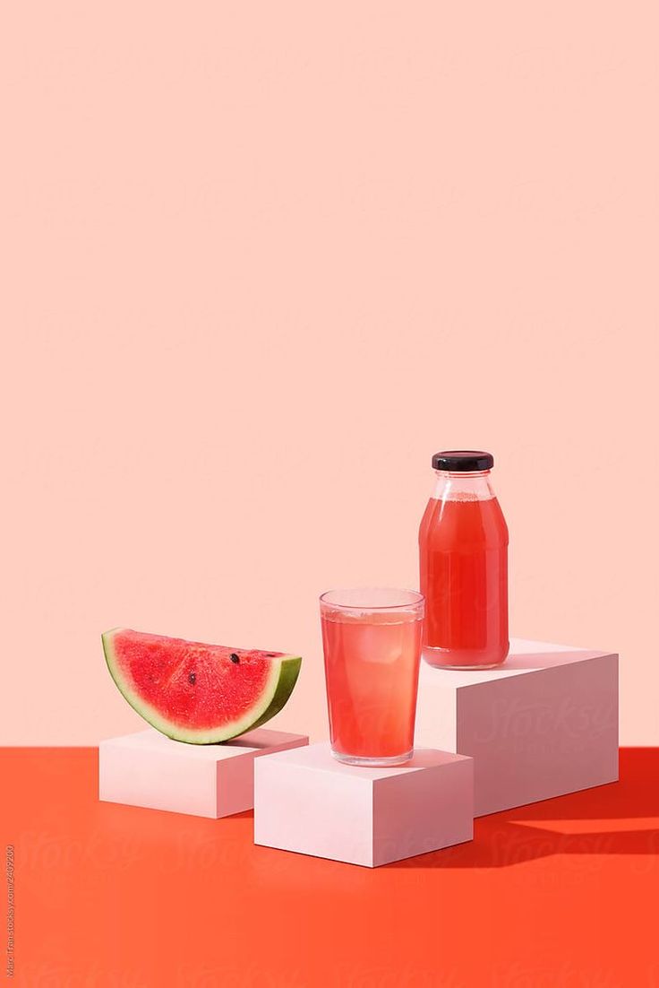 a watermelon and juice are sitting on top of white blocks against a pink background