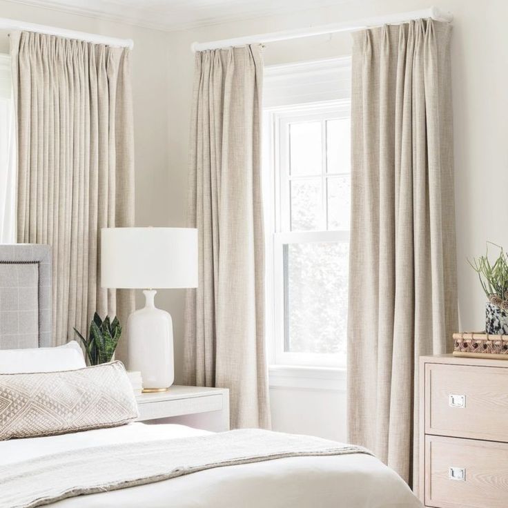 a white bed sitting next to a window in a bedroom under a large window with drapes