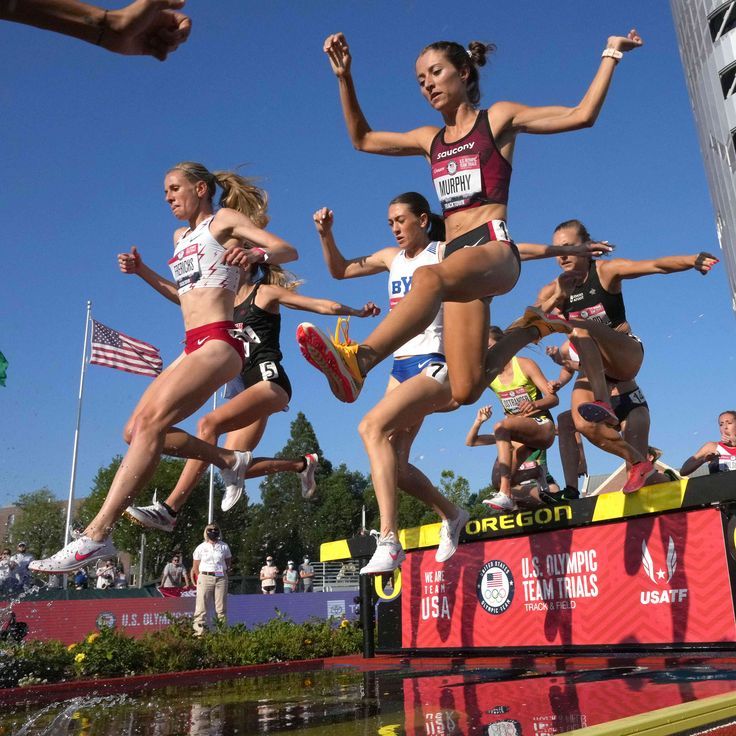 a group of women running in a race on top of a water fountain with an american flag behind them