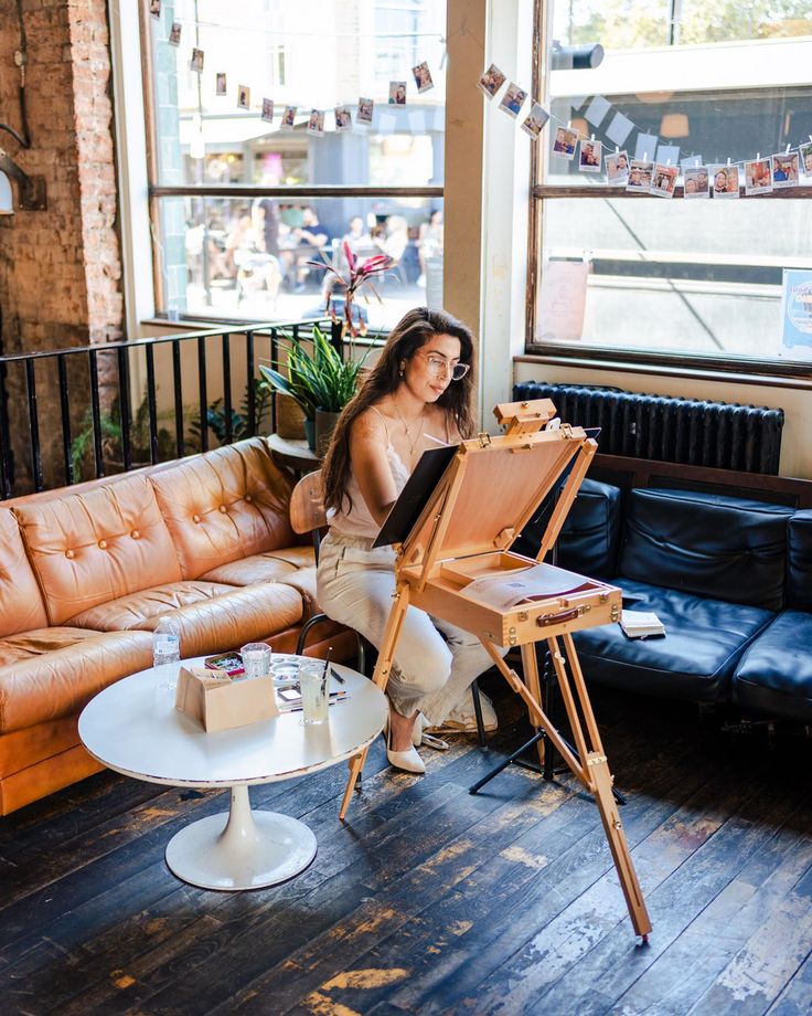 a woman sitting in front of an easel on top of a wooden floor next to a couch