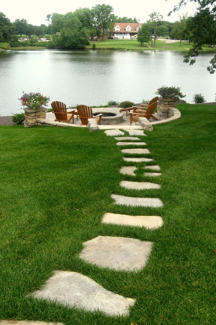 a stone path in the grass leading to a fire pit with chairs on it and a lake in the background