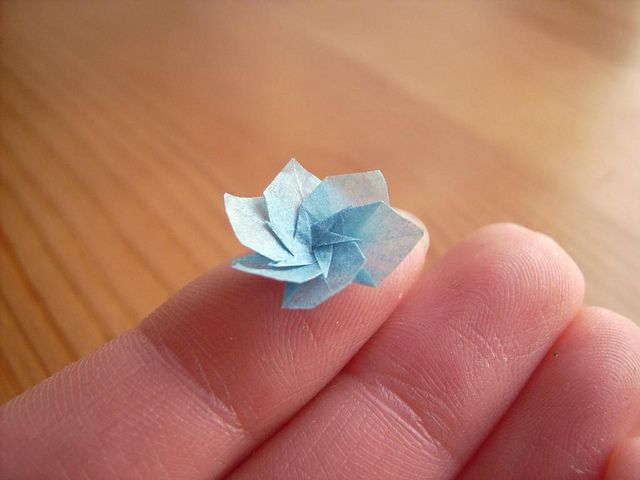 a small blue origami flower sitting on top of a persons finger in front of a wooden table