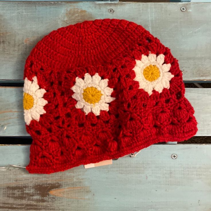 a red crocheted hat with white and yellow flowers on it sitting on a wooden bench