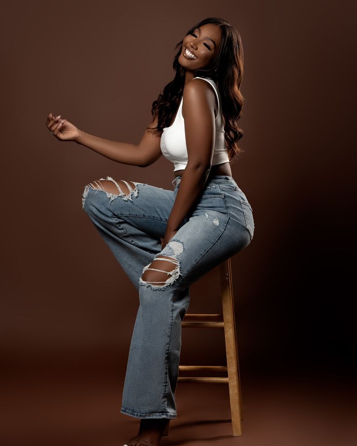 a woman sitting on top of a wooden chair wearing ripped jeans and white tank top