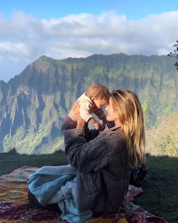 a woman holding a child while sitting on top of a hill with mountains in the background