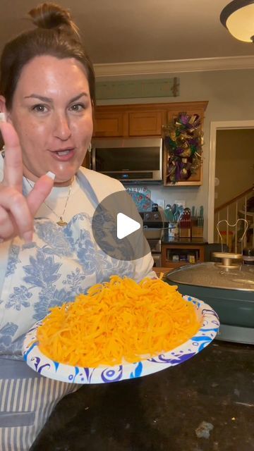 a woman holding up a plate with cheese on it and pointing to the camera while standing in a kitchen