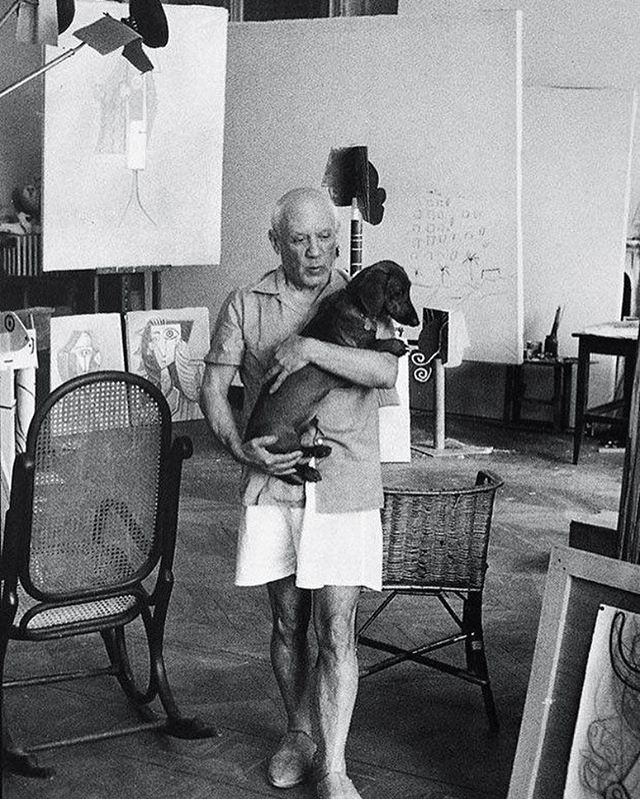 an old man holding a dog in his arms while standing next to chairs and paintings