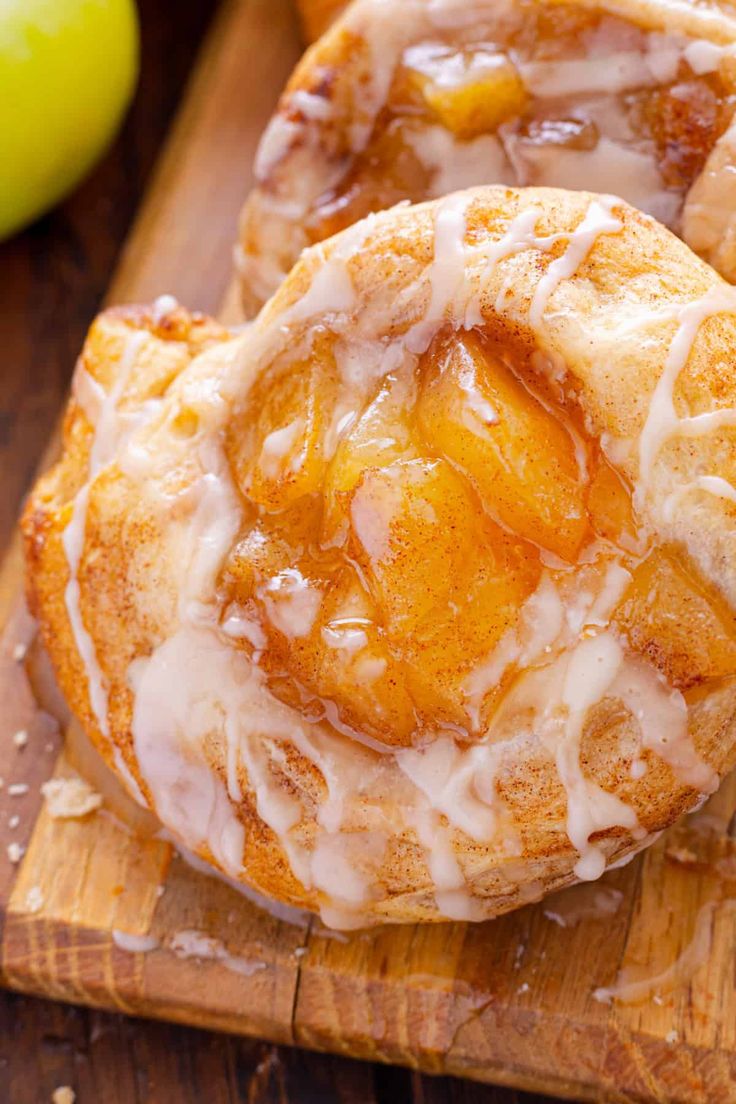 two glazed donuts sitting on top of a wooden cutting board next to an apple