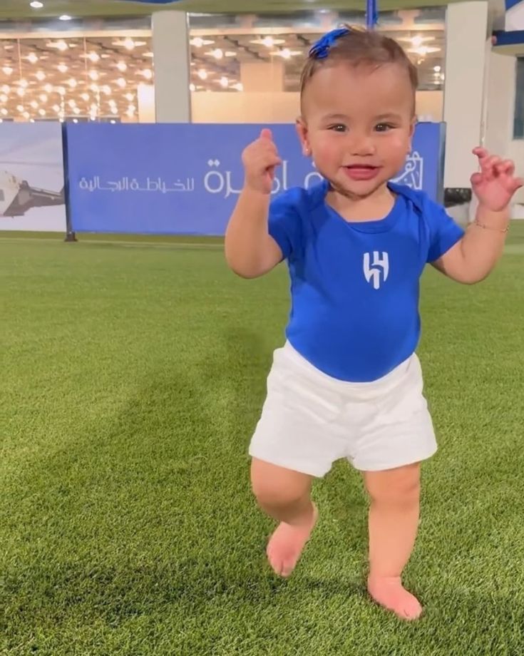 a baby in blue shirt and white shorts standing on grass with one hand raised up