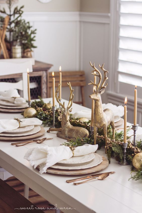 the table is set for christmas dinner with gold and white plates, deer figurines, evergreen greenery, and candlesticks