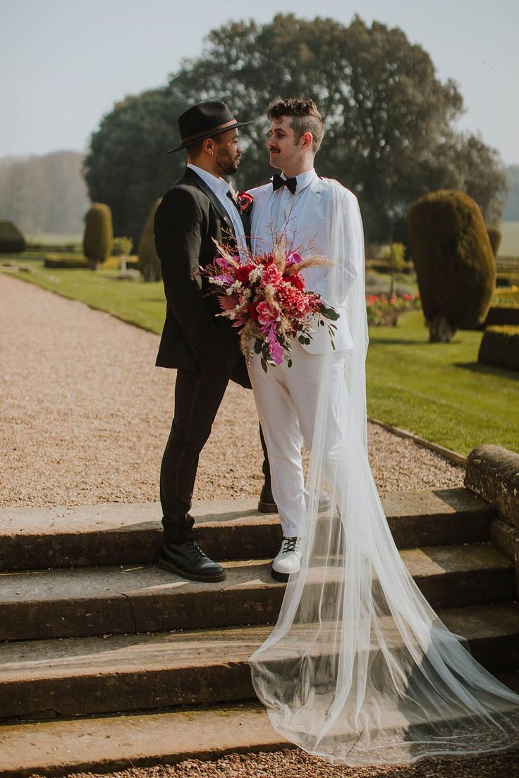 Groom in a black suit and fedora hat standing with his partner at Prestwold Hall Barns in a white groom suit and cape holding a vibrant red bouquet Wedding Suit With Veil, Unconventional Wedding Suit, Mens Wedding Suit With Cape, Wedding Suit With Cape, Fancy Wedding Suit, Enby Wedding, Gay Wedding Outfits, Groom In White Suit, Gay Wedding Suits