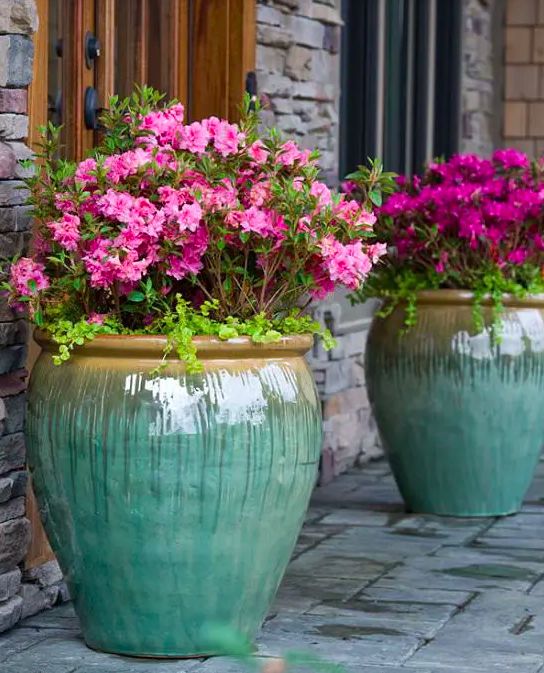 two large green vases filled with pink flowers on a stone walkway next to a brick building