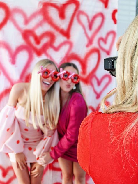 two women are taking pictures in front of a heart - shaped wall with red glasses