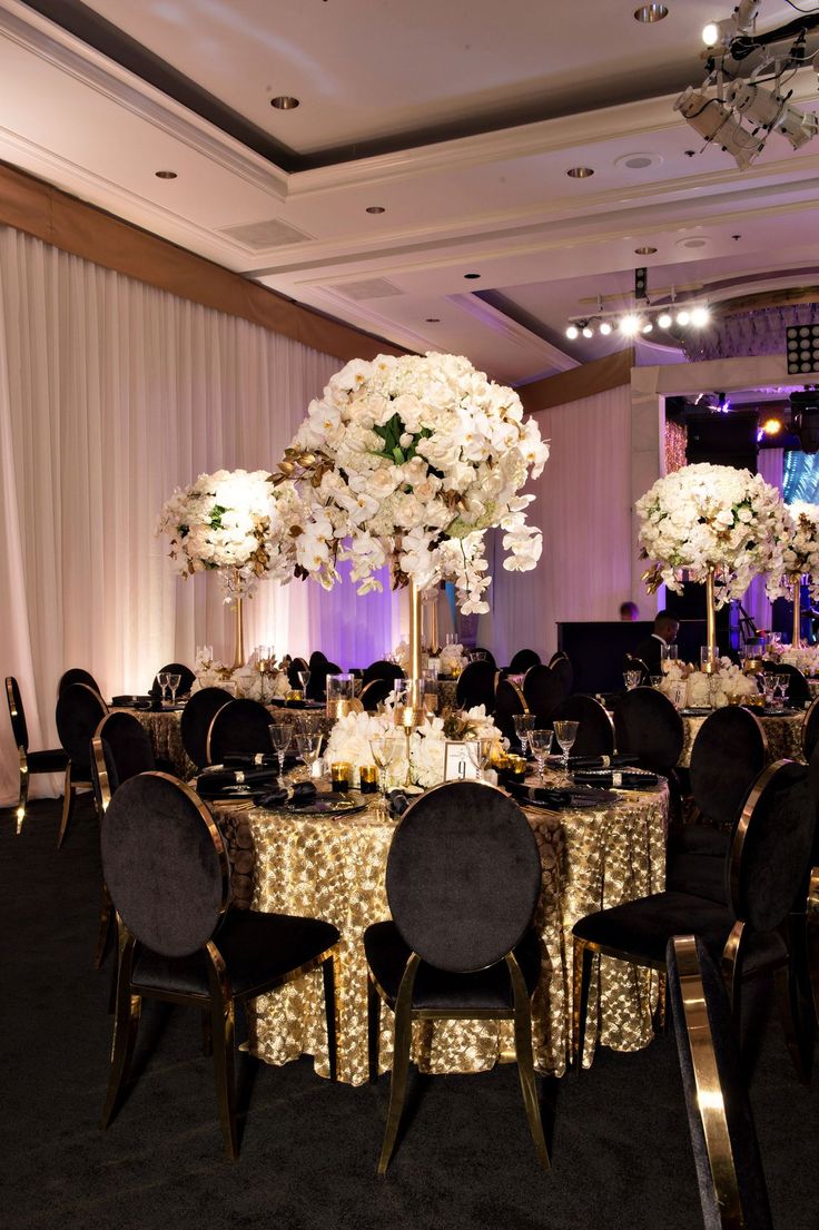 the tables are set up with black chairs and gold tablecloths, white flowers in tall centerpieces