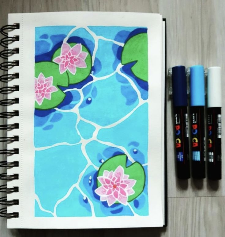 an open notebook with water lilies on it next to three markers and two pens