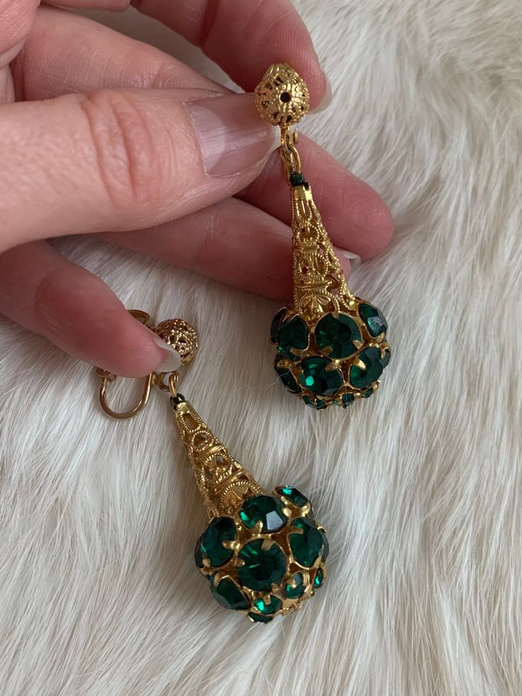 "These are vintage mid-century clip on earrings. They are in a gold tone metal that is bright and shows no signs of wear or tarnish. The base of the earrings have round shape emerald tone rhinestones that form a sphere. The earrings are in excellent condition. Measurements: Length: 2\" | Width: .75\" Unless otherwise stated all vintage items are used and may have minor to moderate wear or discoloration considering the age of the item. Most items have been hand washed, spot cleaned, or profession Brass Dangle Clip-on Earrings, Gold Jeweled Evening Clip-on Earrings, Vintage Green Jeweled Earrings, Gold Costume Jewelry Earrings For Wedding, Evening Metal Clip-on Crystal Earrings, Formal Jeweled Metal Clip-on Earrings, Metal Clip-on Crystal Earrings For Evening, Antique Gold Jeweled Earrings, Antique Gold Dangle Clip-on Earrings