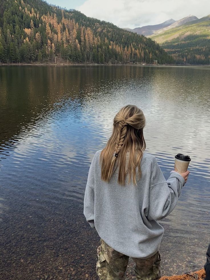 a woman standing on the shore of a lake holding a coffee cup in her hand