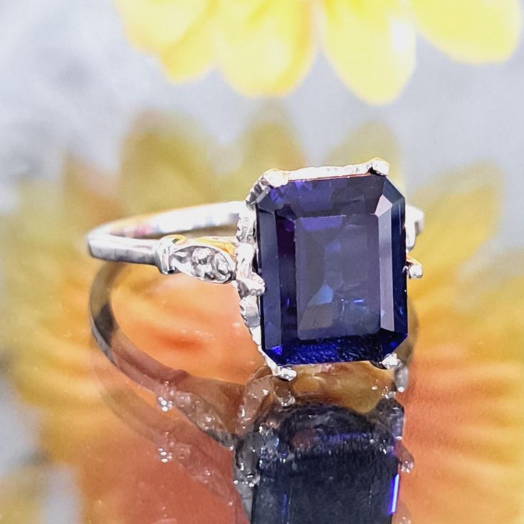 an image of a ring with a blue stone on it and flowers in the background